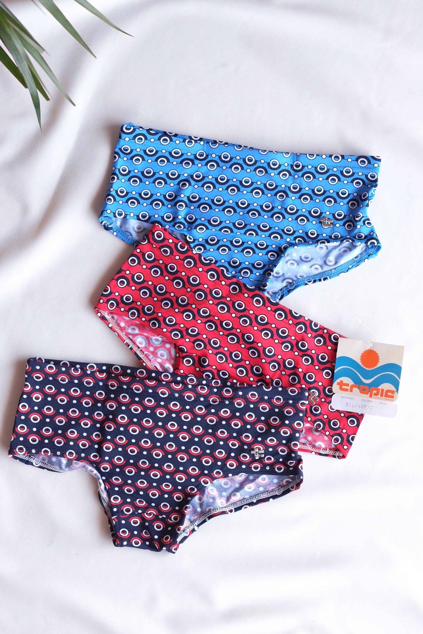 Three pairs of vintage TROPIC swim briefs stacked on top of each other. The briefs are in different colors, including blue,
 red, and navy, and feature a repeating circular pattern.