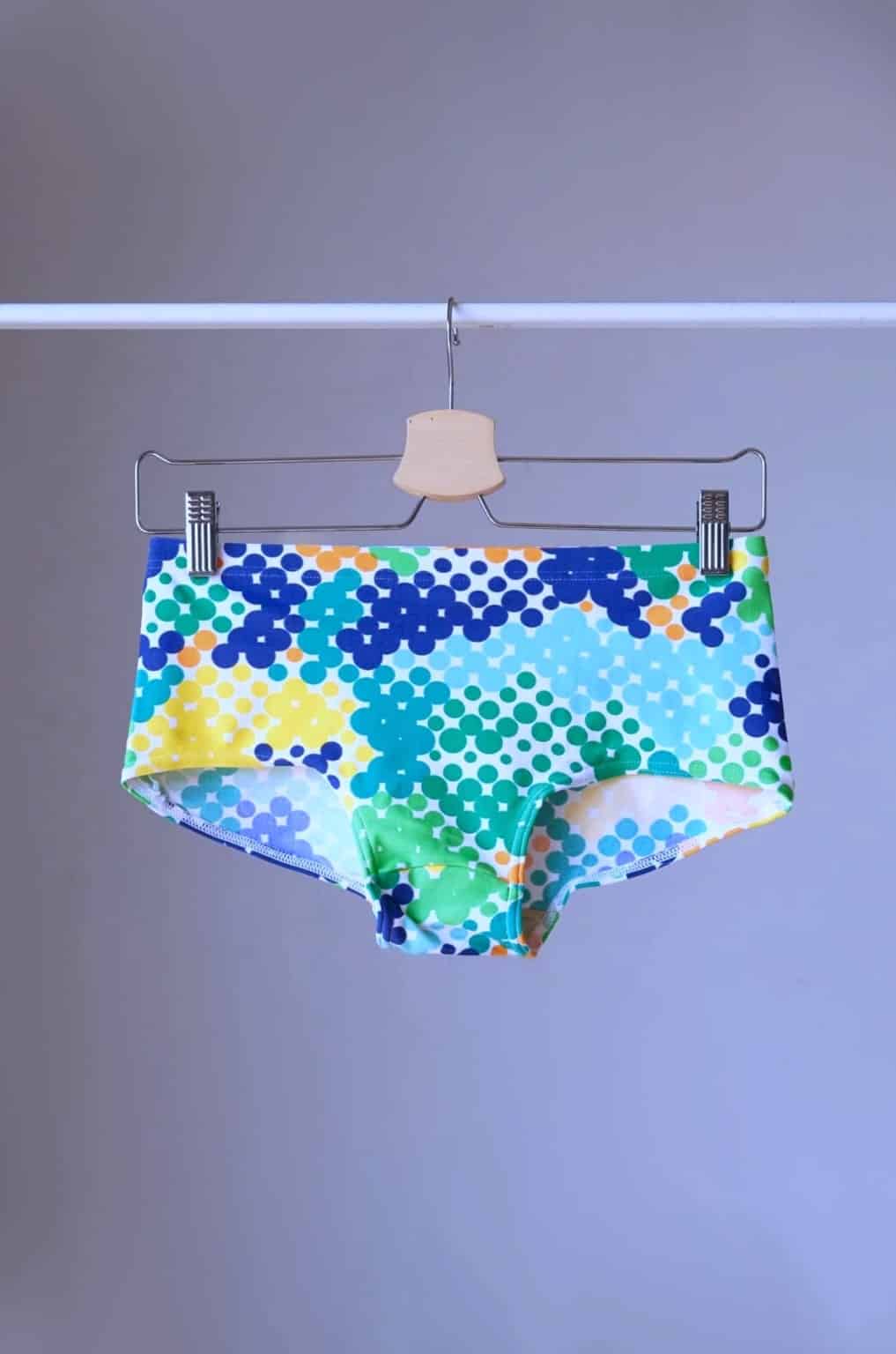 TROPIC Vintage Retro Dot Print Swim Briefs on a hanger, showcasing the blue-green variant with a vibrant geometric dotted design. The pattern includes shades of green, blue, yellow, and orange dots for a bold, retro look.