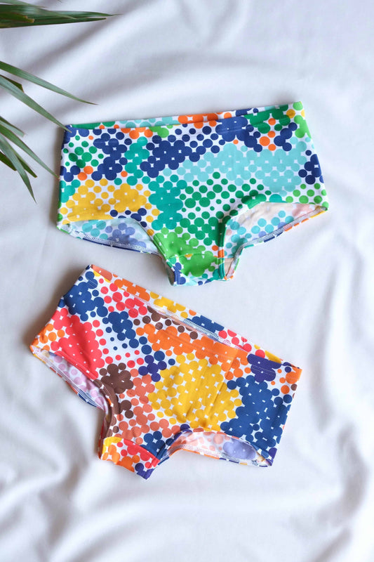 Two pairs of Tropic colorful swim briefs with a retro dotted pattern in vibrant colors, including blues, greens, yellows, and oranges, are laid out on a white surface. One pair has a lighter color scheme, while the other has a more bold and contrasting palette.