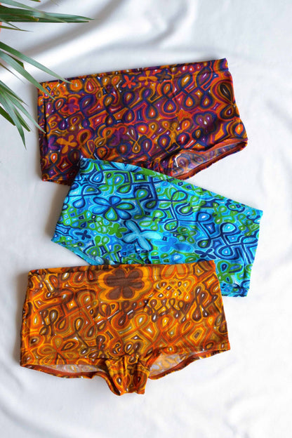 Three pairs of vintage 70s psychedelic men's swim briefs in vibrant colors—red, blue, and orange—laid out on a white surface next to green palm leaves.