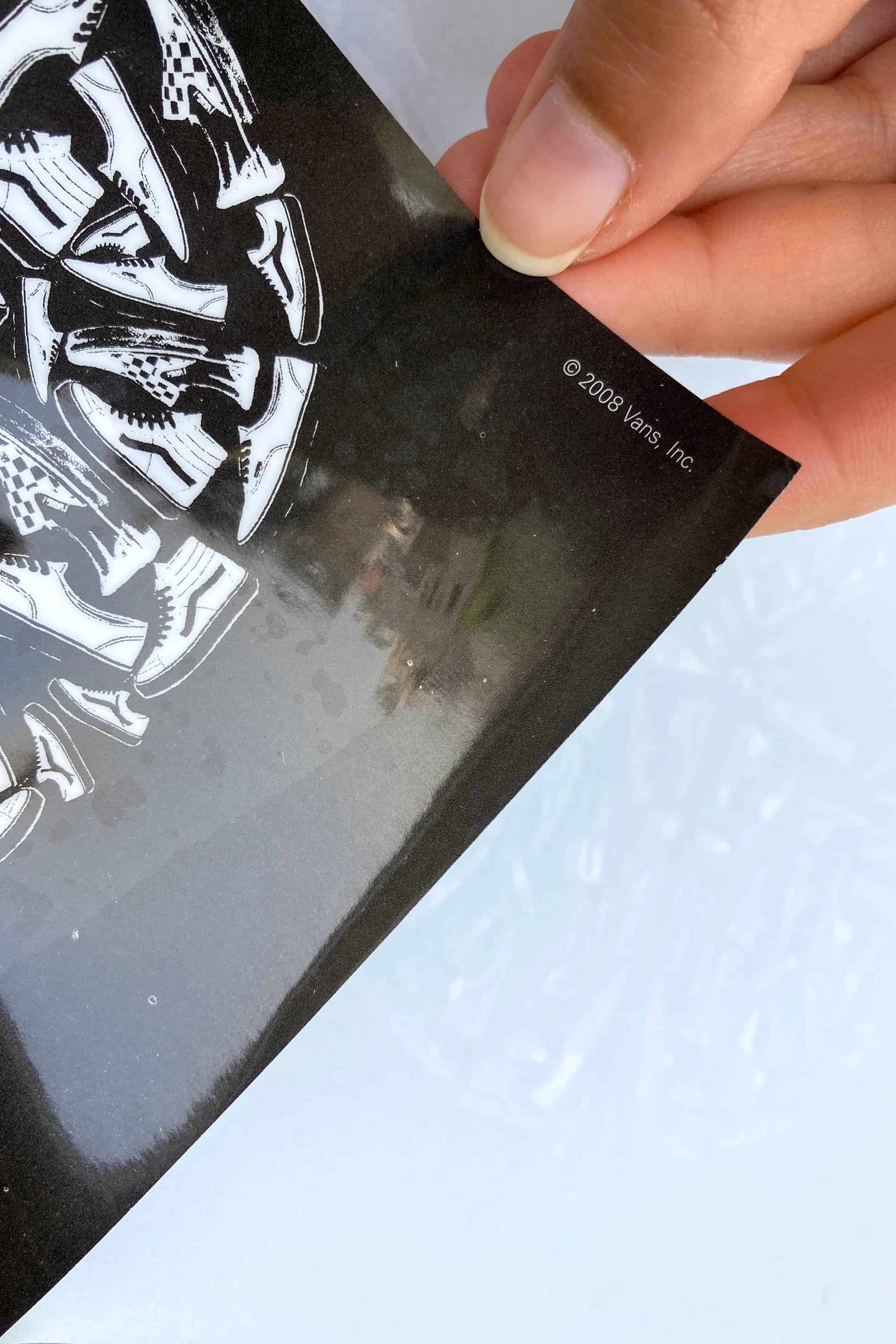 Detail shot of the corner of the Vans window cling showing sneaker graphics and © 2008 Vans, Inc. marking, held by hand against a backlit surface.