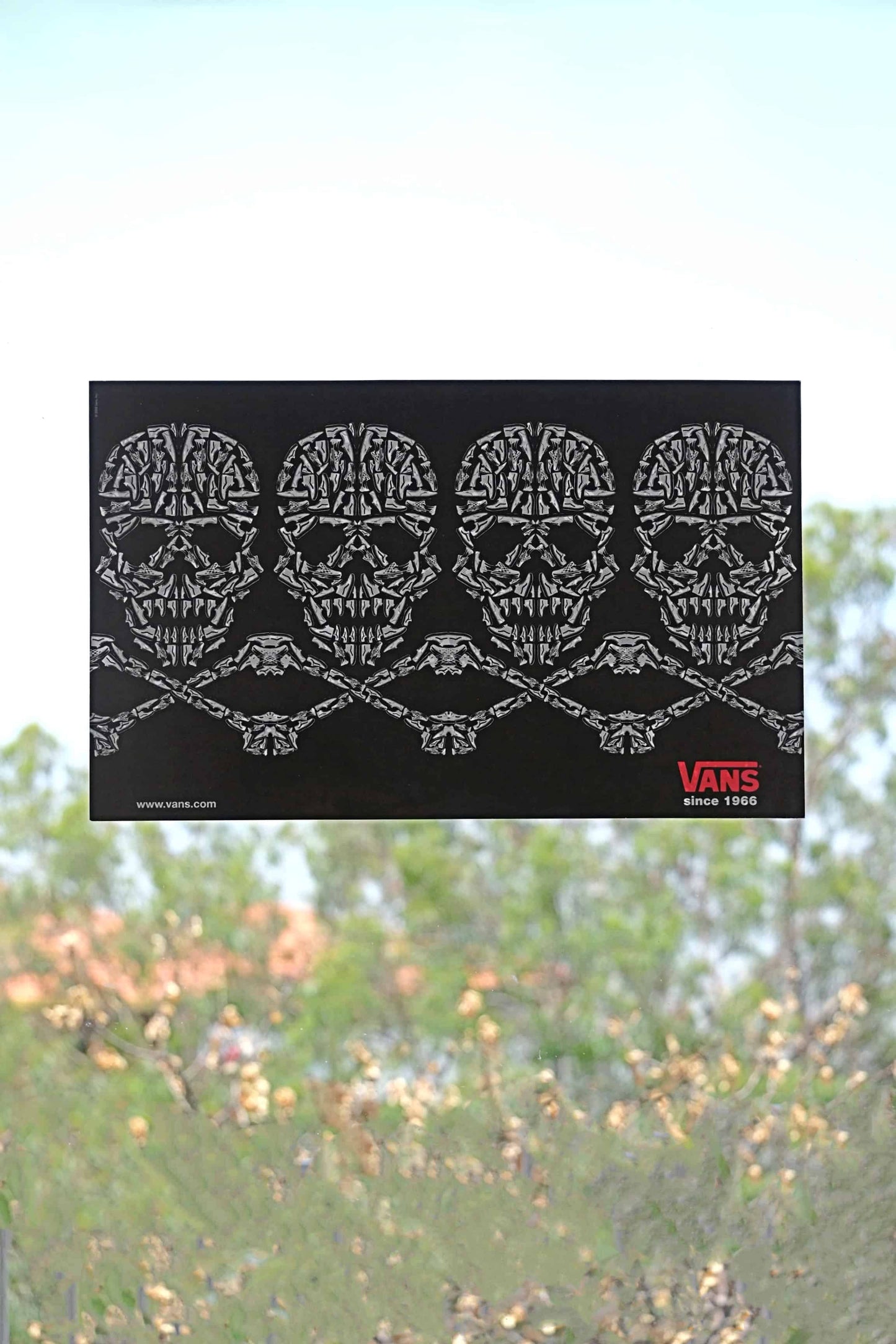 Front view of Vans 2008 static cling window sticker with skulls and crossbones design, made from sneaker illustrations, placed on a clear window with natural light and trees behind.