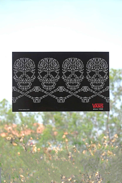 Front view of Vans 2008 static cling window sticker with skulls and crossbones design, made from sneaker illustrations, placed on a clear window with natural light and trees behind.