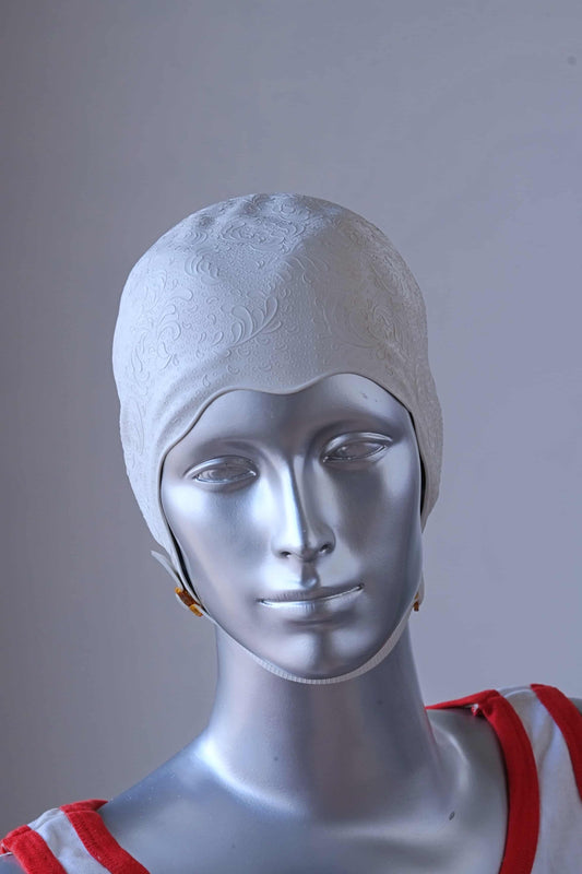 A vintage 1960s white swim cap made of textured rubber with an embossed waves pattern, worn by a silver mannequin. The cap features a secure chin strap with metal clasps. The mannequin is set against a neutral background.