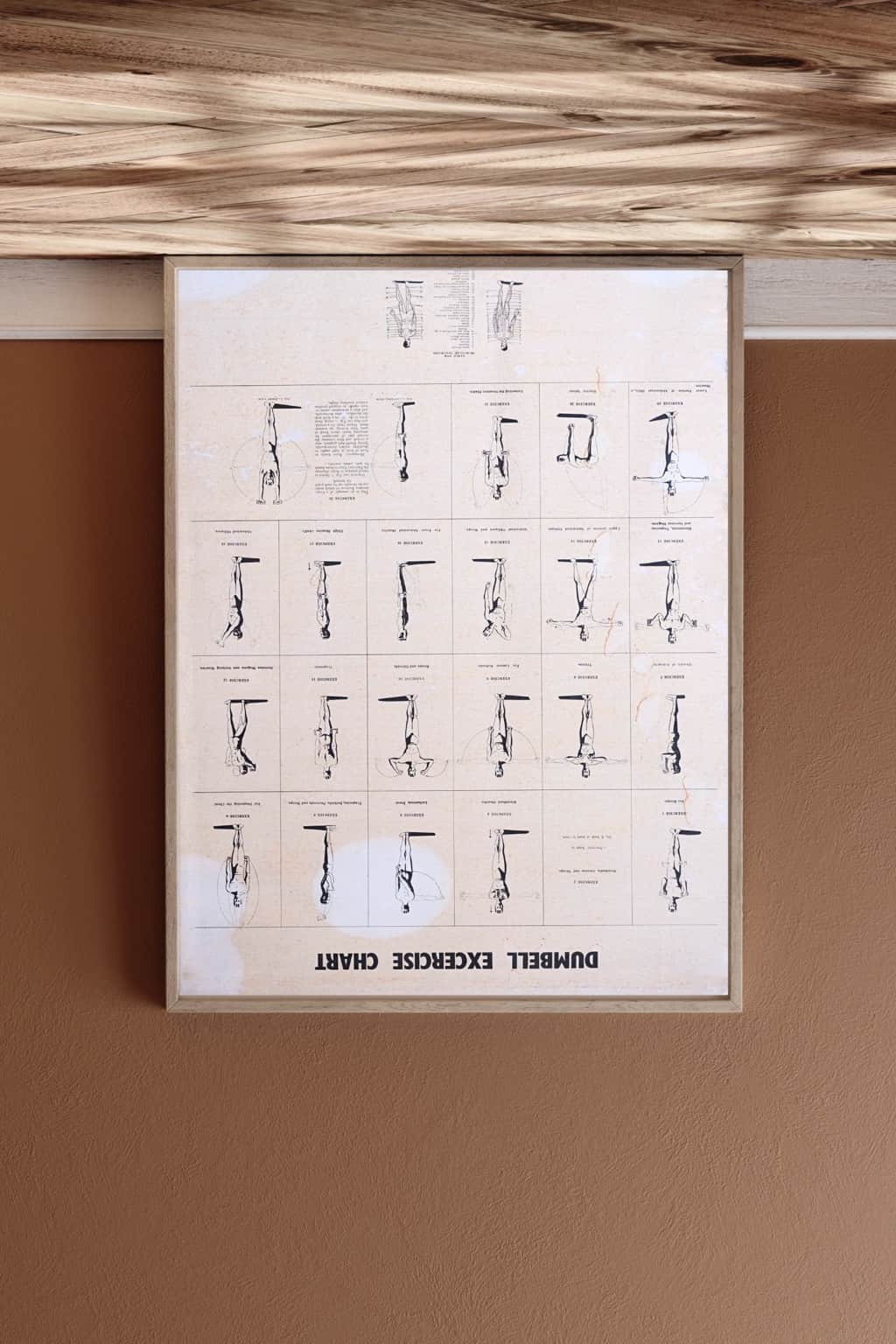Vintage dumbbell workout chart from the 1970s in a brown frame, styled against a tan wall and wood floor.