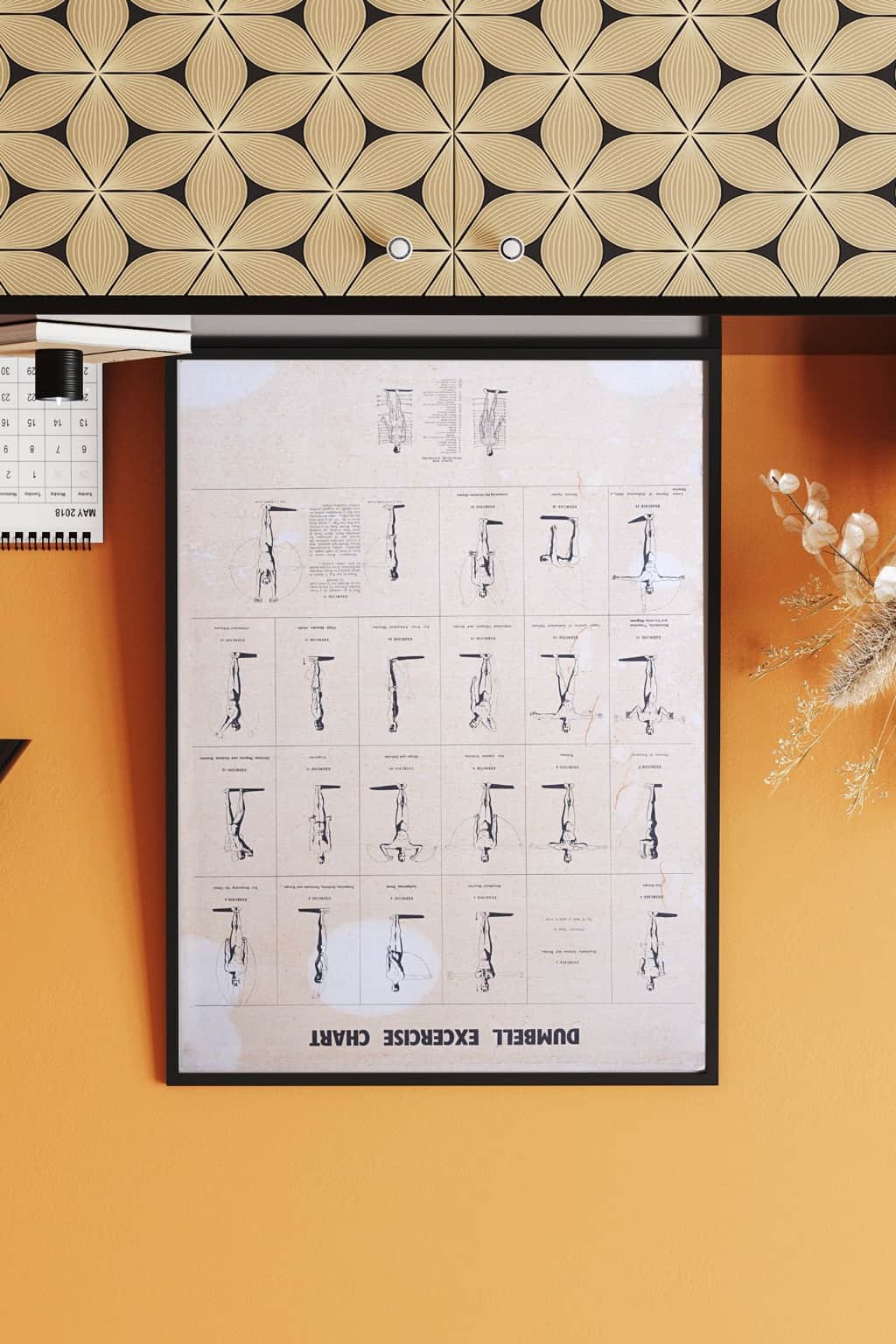 Framed 1970s dumbbell workout chart with orange-yellow wall background, styled with books and dried florals.