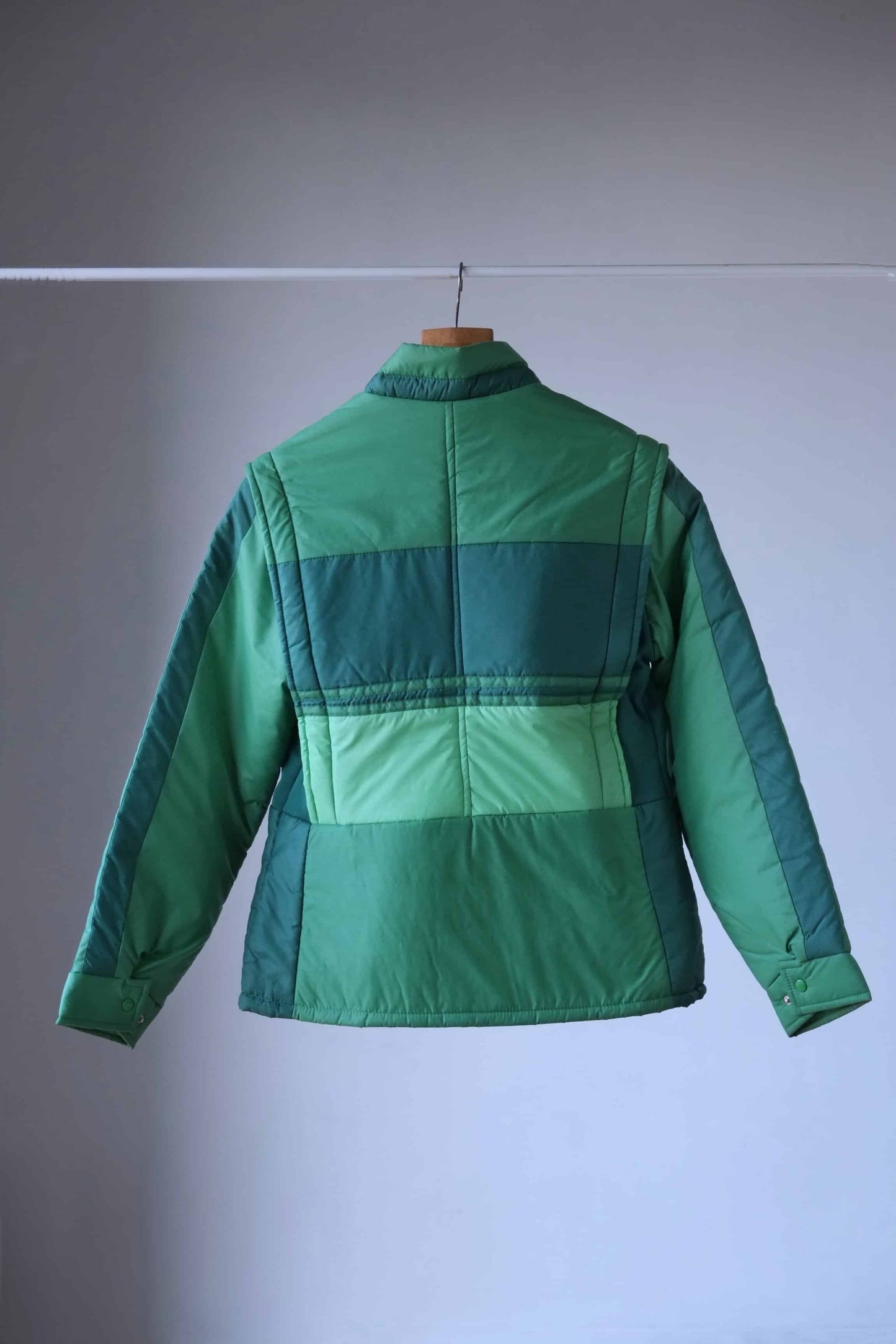 Back view of a vintage 80s green puffer jacket with a retro colorblock design, featuring various shades of green. The jacket has a full zip closure, high collar, and zippered pockets, and is displayed on a wooden hanger against a neutral gray background.