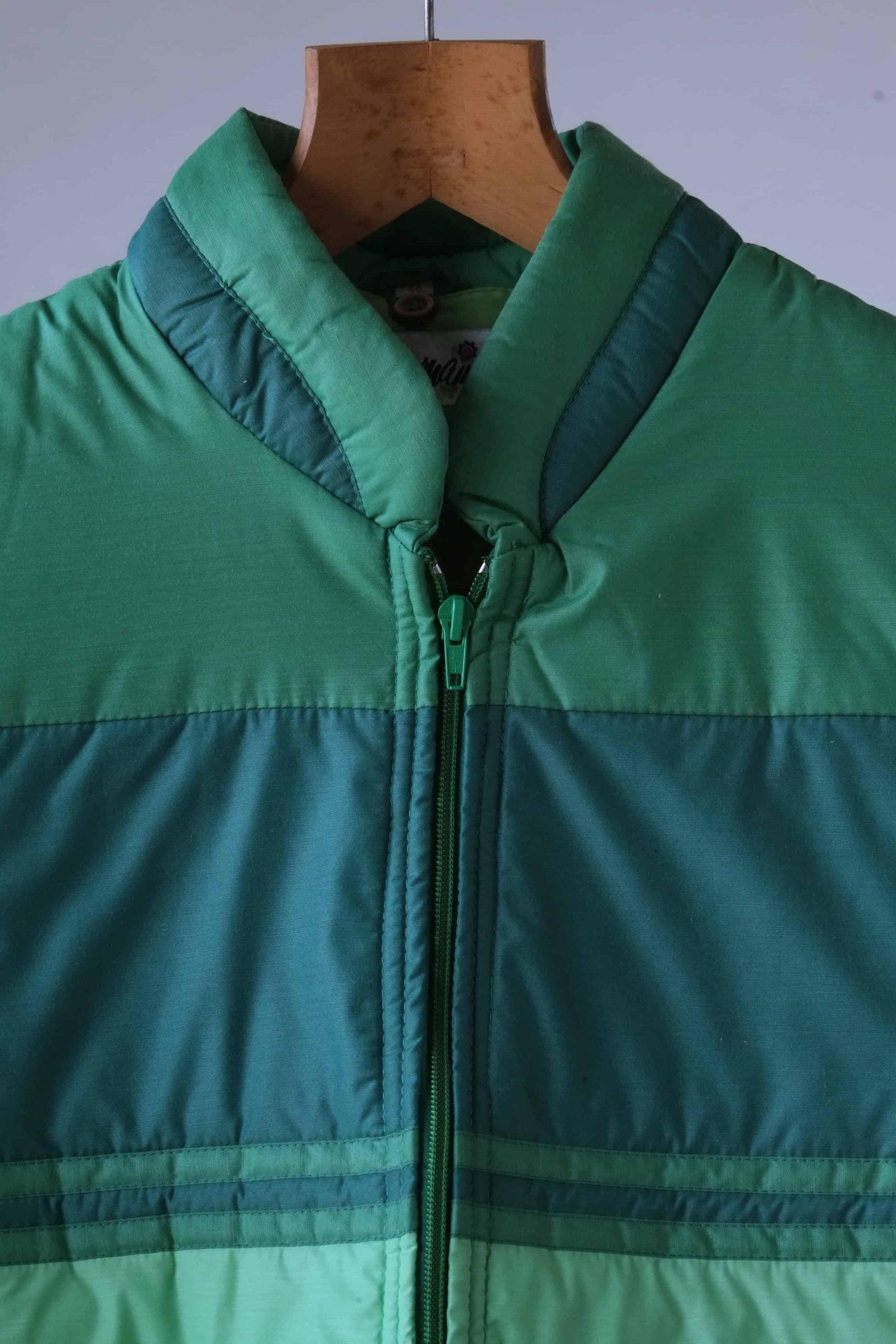 Close-up of a vintage 80s green puffer jacket, highlighting the retro colorblock design, high collar, and front zipper. The fabric texture and stitching details are visible, showcasing the jacket’s quality and construction. The jacket is displayed on a wooden hanger against a neutral gray background.