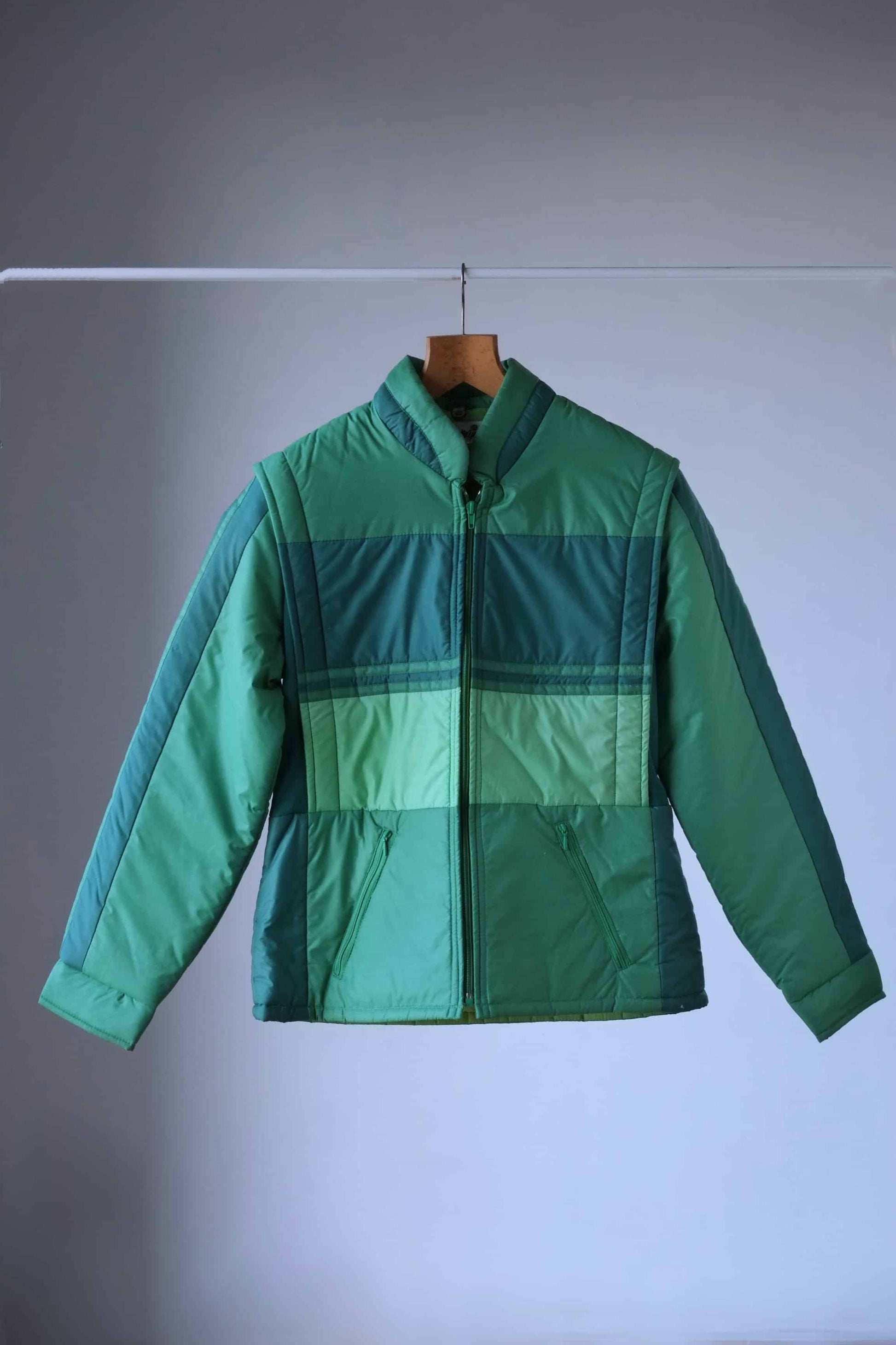 A vintage 80s green puffer jacket with a retro colorblock design, featuring various shades of green. The jacket has a full zip closure, high collar, and zippered pockets, and is displayed on a wooden hanger against a neutral gray background.
