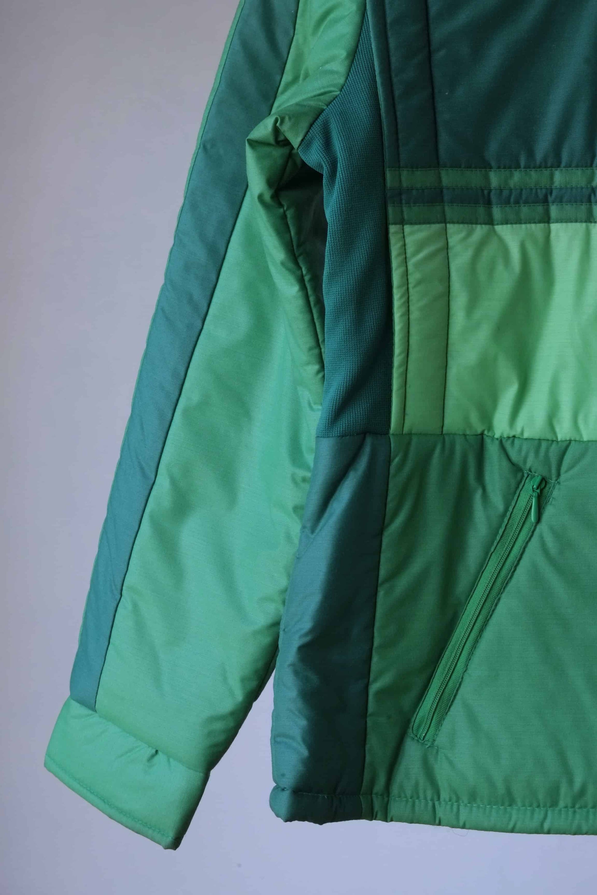 Close-up of the sleeve and side details of a vintage 80s green puffer jacket, showcasing the retro colorblock design, quilted padding, and zippered pocket. The stitching, fabric texture, and cuff structure are visible.