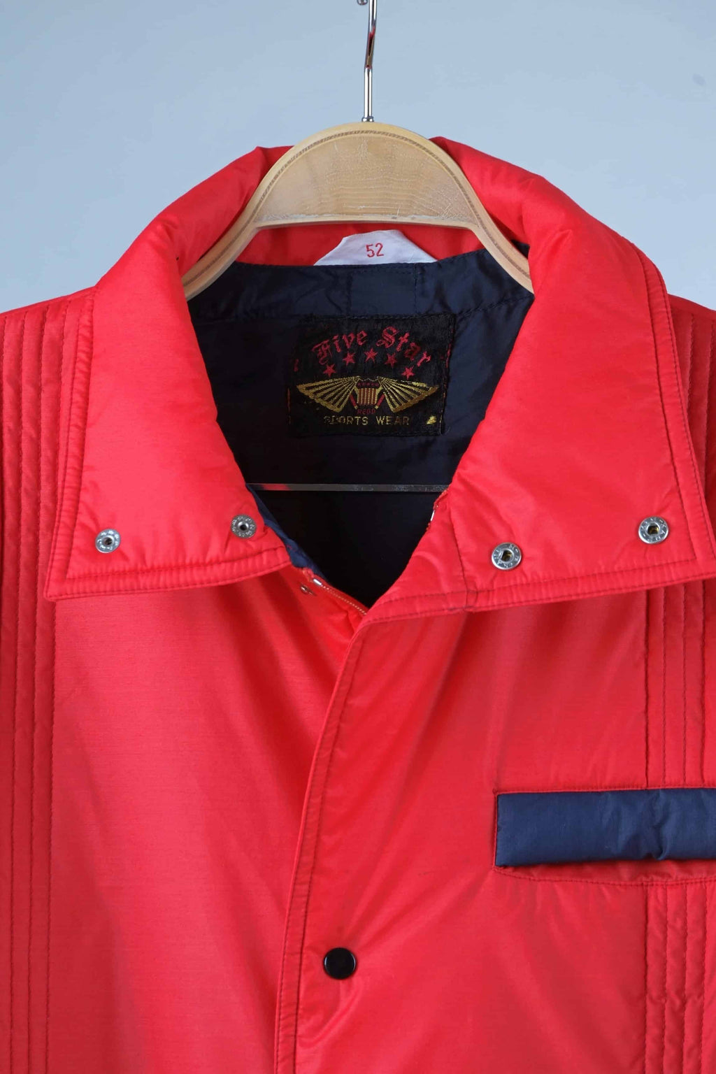 Close view of the collar of a Men's red vintage 80s puffer ski jacket with blue pocket details, a stand-up collar, and a zipper/snap button closure, displayed on a hanger.