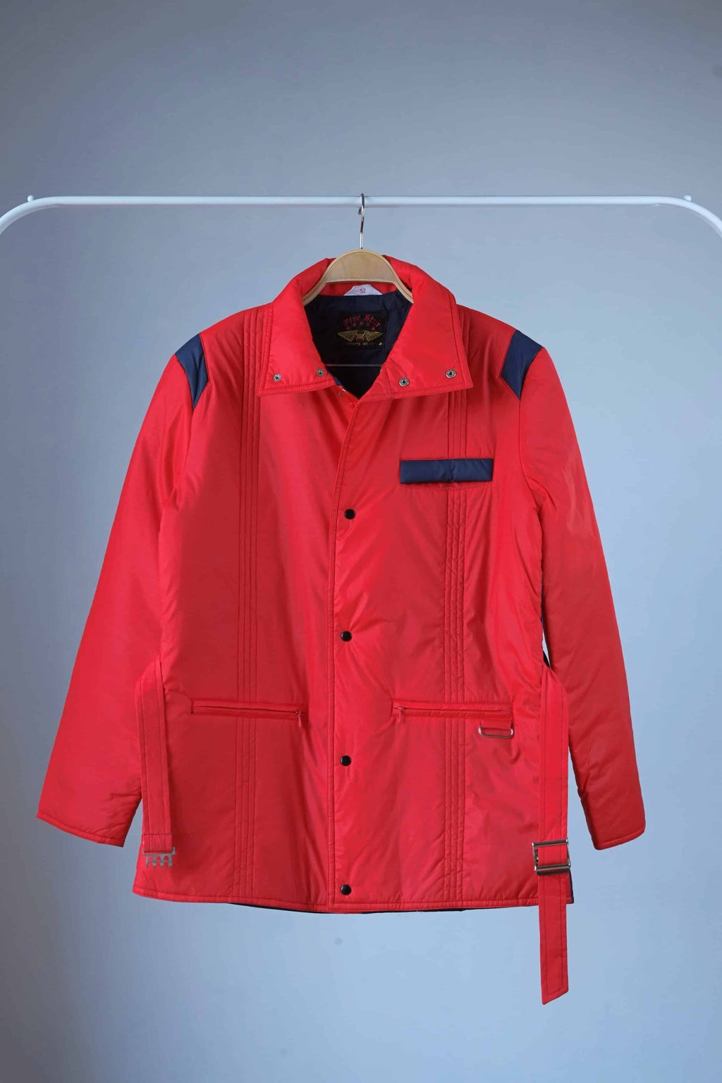 Men's red vintage 80s puffer ski jacket with a belted waist, blue shoulder bands and pocket details, a stand-up collar, and a zipper/snap button closure, displayed on a hanger.