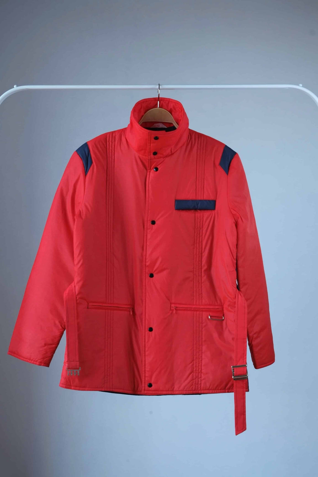 Men's red vintage 80s puffer ski jacket with a belted waist, blue shoulder bands and pocket details, shown with a closed stand-up collar, and a zipper/snap button closure, displayed on a hanger.