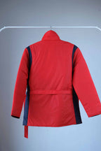 Back view of a Men's red vintage 80s puffer ski jacket with a belted waist, blue shoulder bands and pocket details, a stand-up collar, and a zipper/snap button closure, displayed on a hanger.