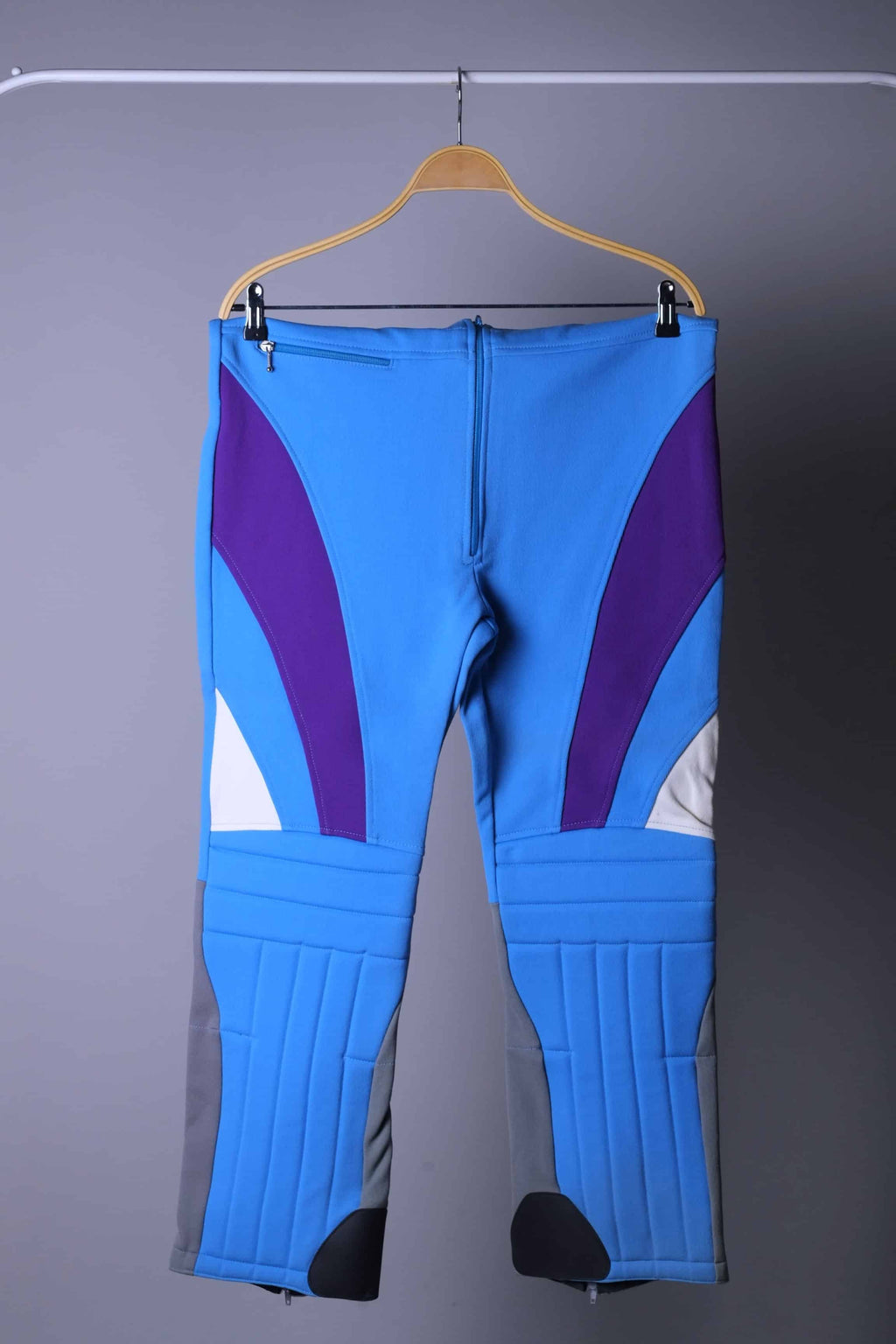 A pair of vintage 90s high-waisted slalom ski bibs in blue, purple, and white color-block, displayed folded on a hanger.