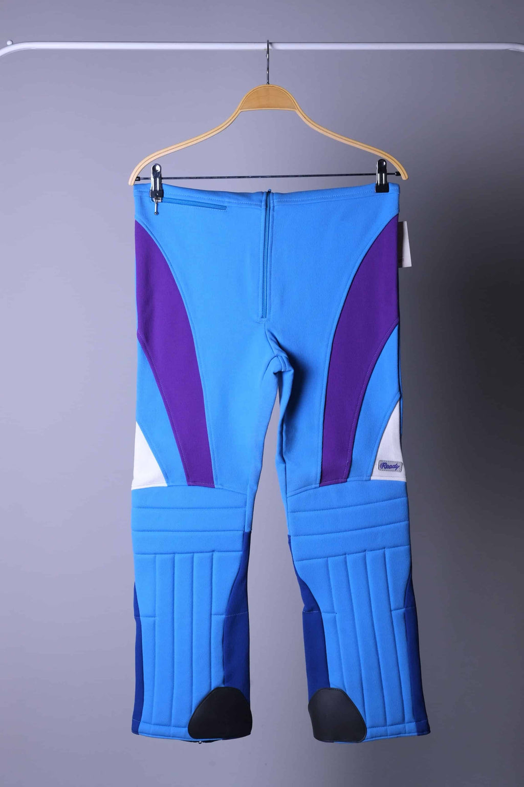A pair of vintage 90s high-waisted slalom ski bibs in blue, purple, and navy color-block, displayed folded on a hanger.