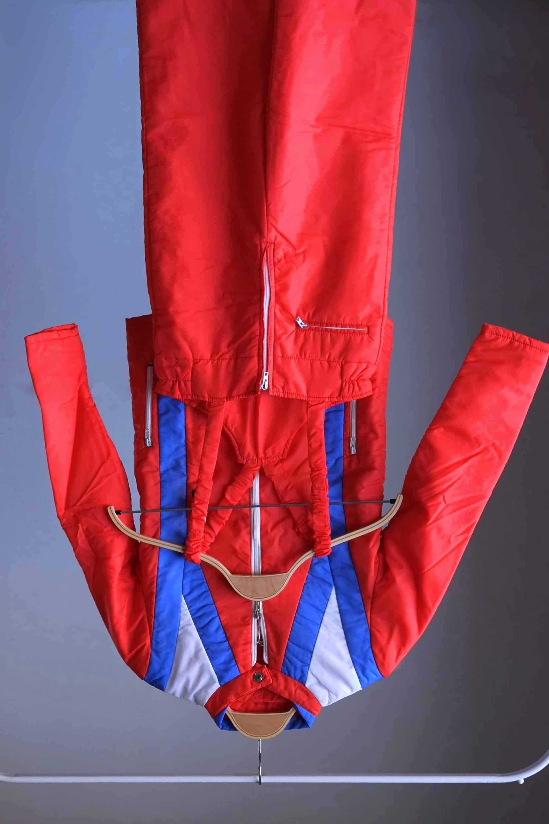 Full view of a two-piece retro ski suit by Warmia featuring a vibrant red insulated jacket with blue geometric details and high-waisted red bib overalls with ruffled elastic straps.
