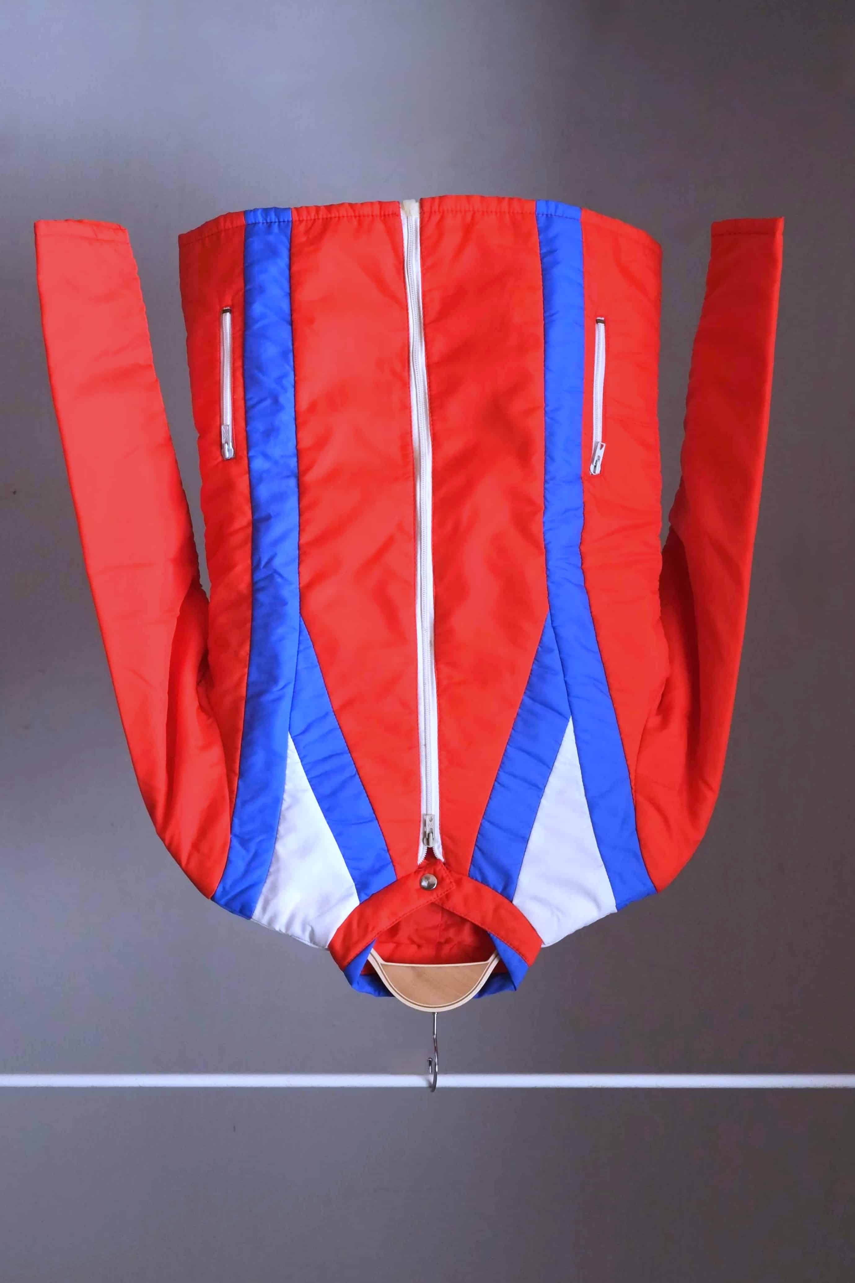 Full view of the jacket from a two-piece retro ski suit by Warmia featuring a vibrant red insulated jacket with blue geometric details.