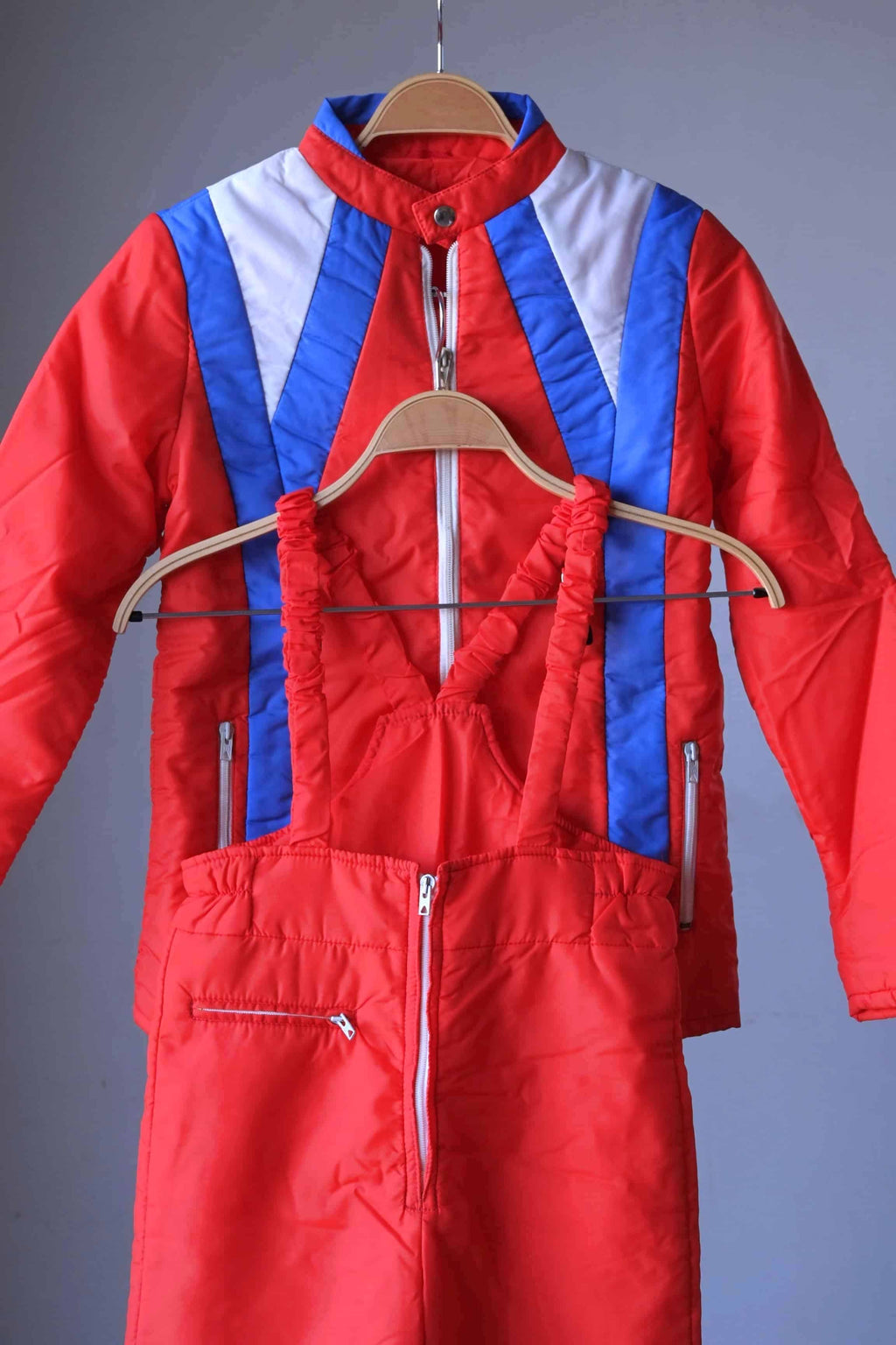 Tight shot of a two-piece retro ski suit by Warmia featuring a vibrant red insulated jacket with blue geometric details and high-waisted red bib overalls with ruffled elastic straps.