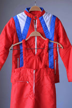 Tight shot of a two-piece retro ski suit by Warmia featuring a vibrant red insulated jacket with blue geometric details and high-waisted red bib overalls with ruffled elastic straps.