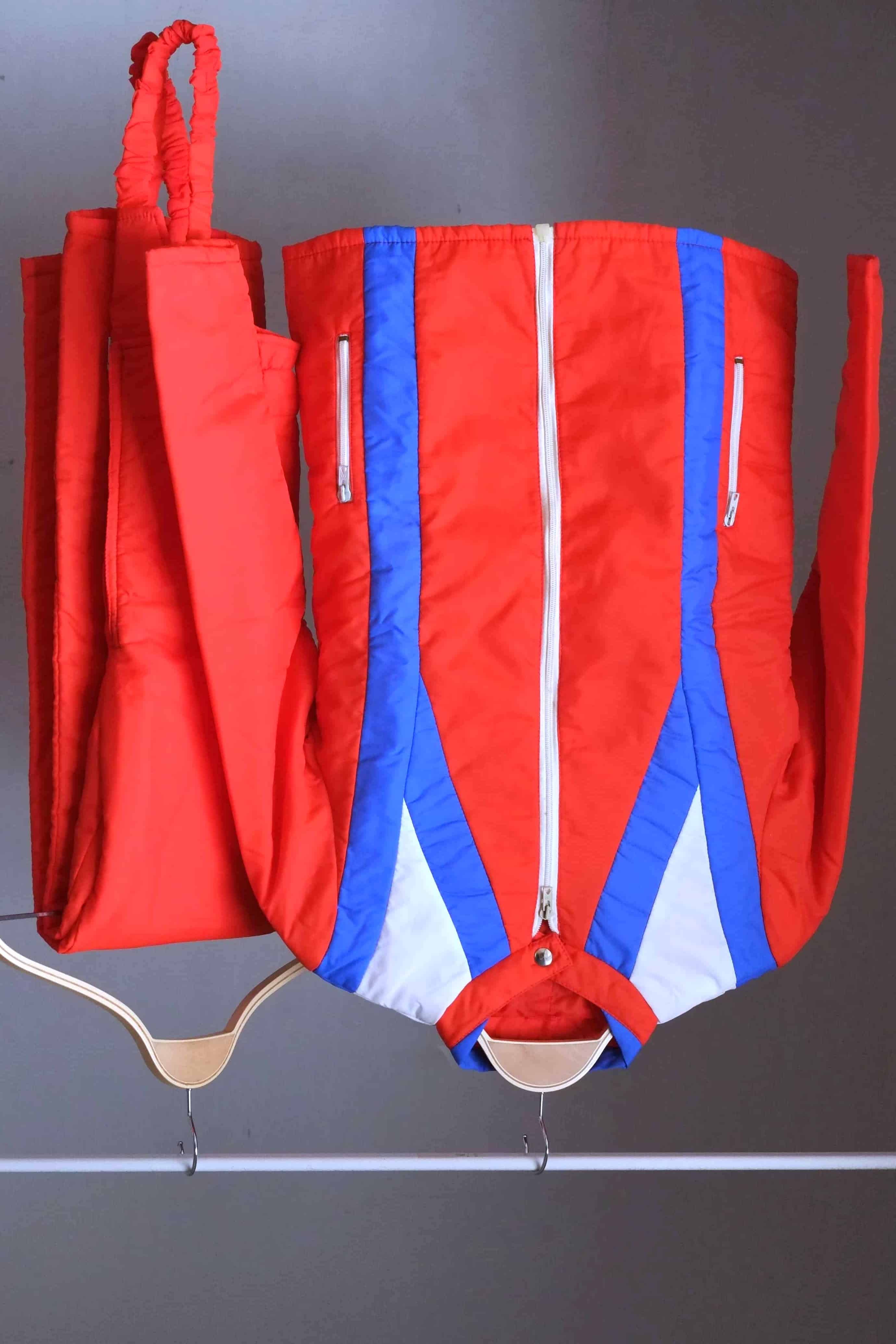 A vintage 70s Warmia red quilted ski jacket with white and blue chevron paneling on the chest, displayed on a wooden hanger alongside matching red bib pants.