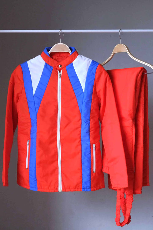 A vintage 70s Warmia red quilted ski jacket with white and blue chevron paneling on the chest, displayed on a wooden hanger alongside matching red bib pants.