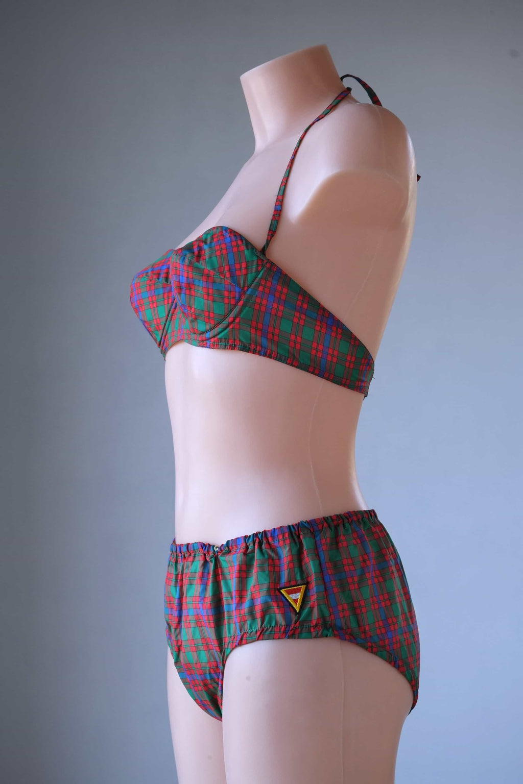 Side profile view of a vintage red and green plaid bikini set on a mannequin, showcasing the cup shape, halter tie, and side hip coverage.