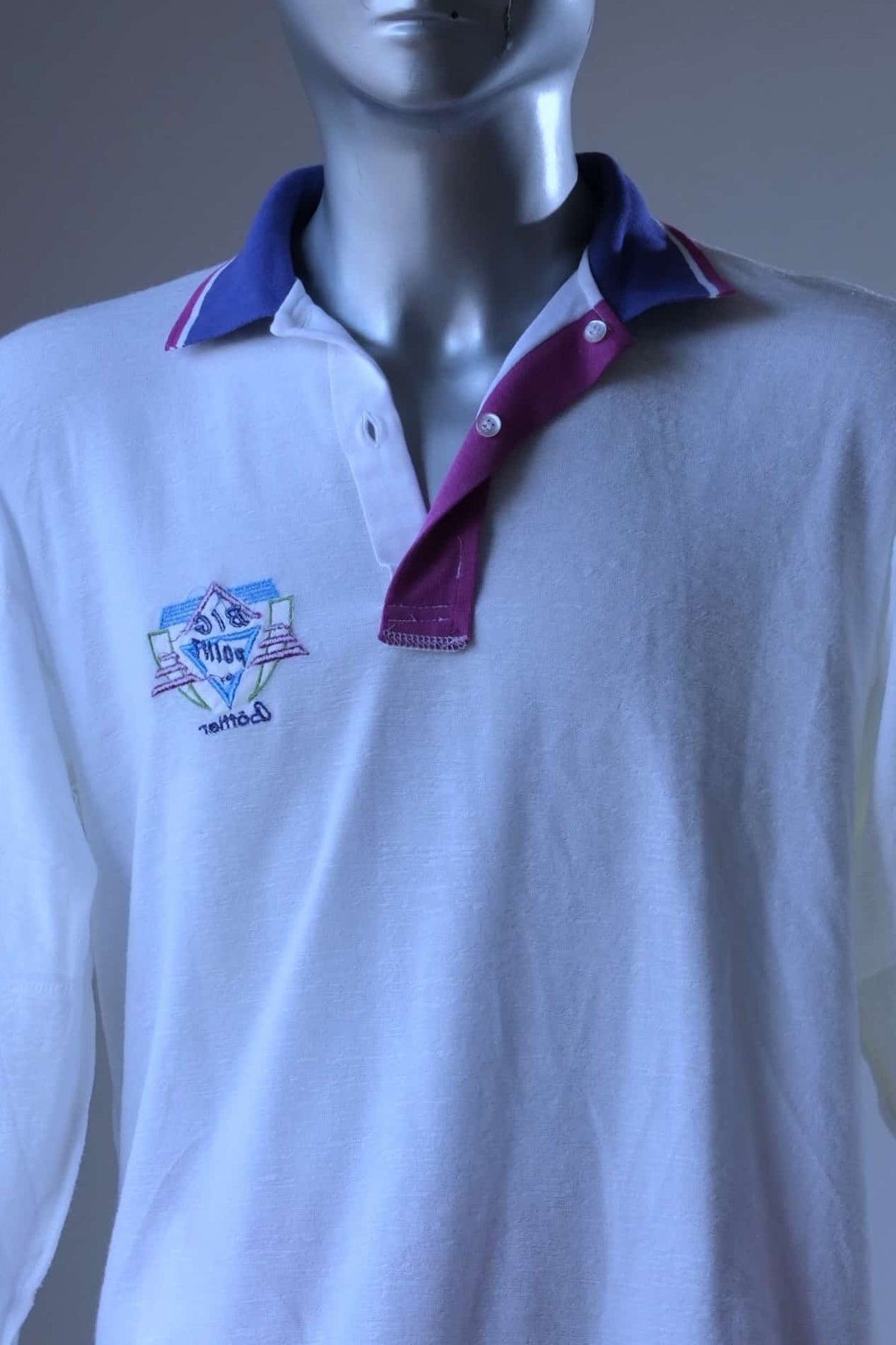 Inside-out view of white Löffler 90s long-sleeve tennis polo on a mannequin, showing the reverse side of the embroidered Big Point logo and the magenta placket facing