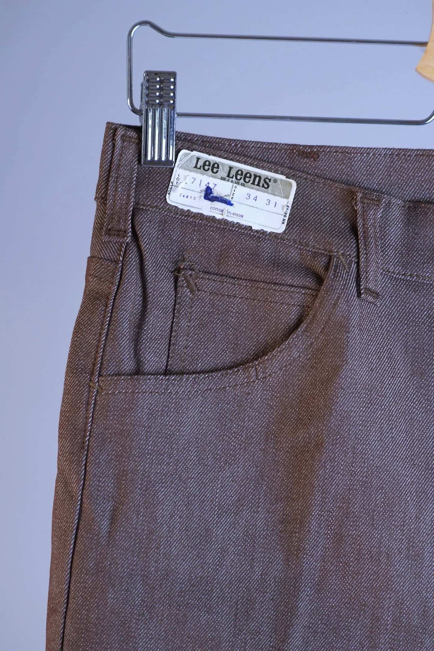 Close-up of the waistband and front pocket of the heather brown Lee Leens, showing the original paper size tag 34x31 and the 'Lee Leens' cloth label.