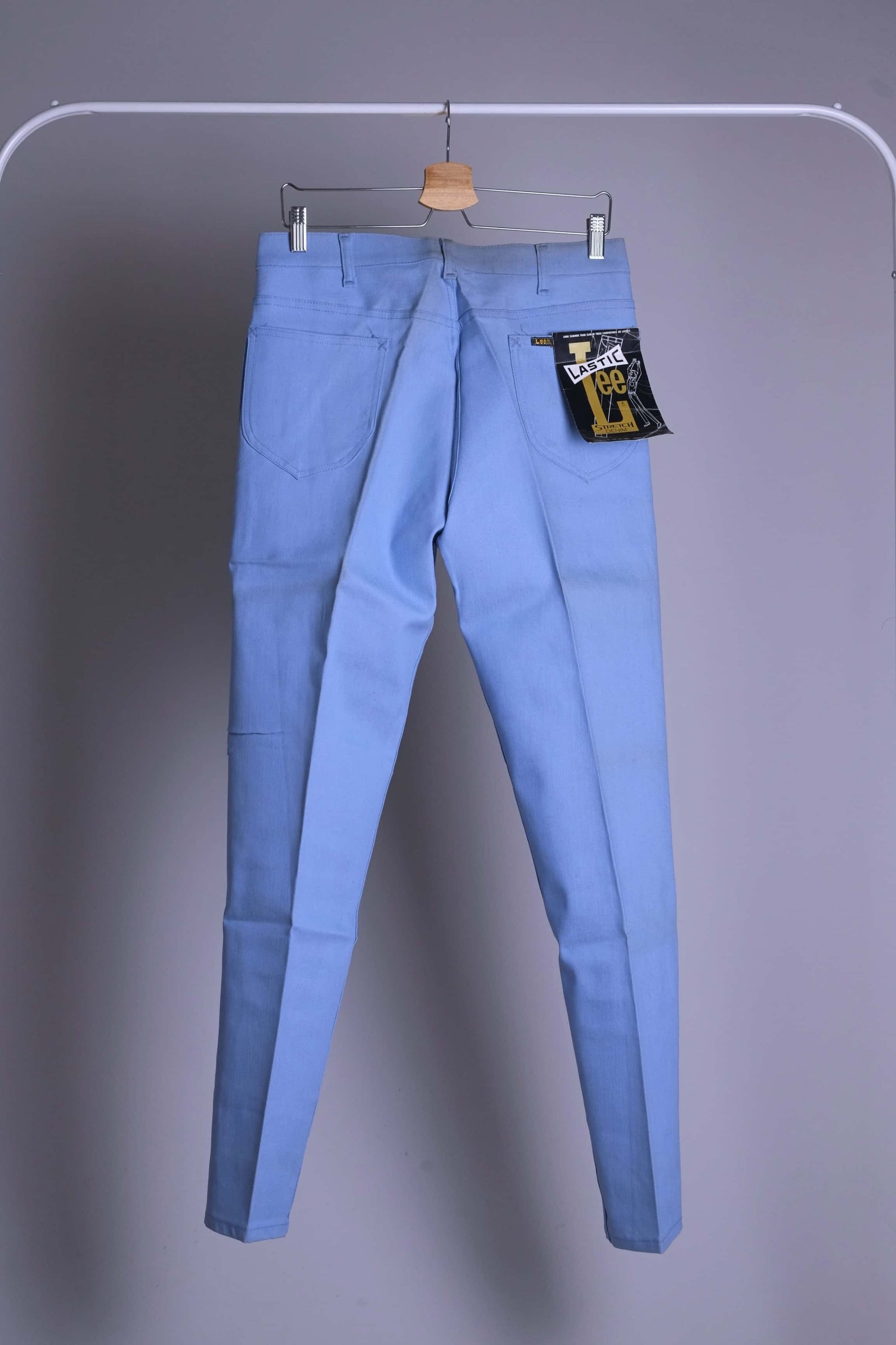 Back view of NOS 70s Lee Leens Lastic Stretch denim trousers in light sky blue, shown hanging on a rack with original black and yellow 'Lastic Lee' paper tag.