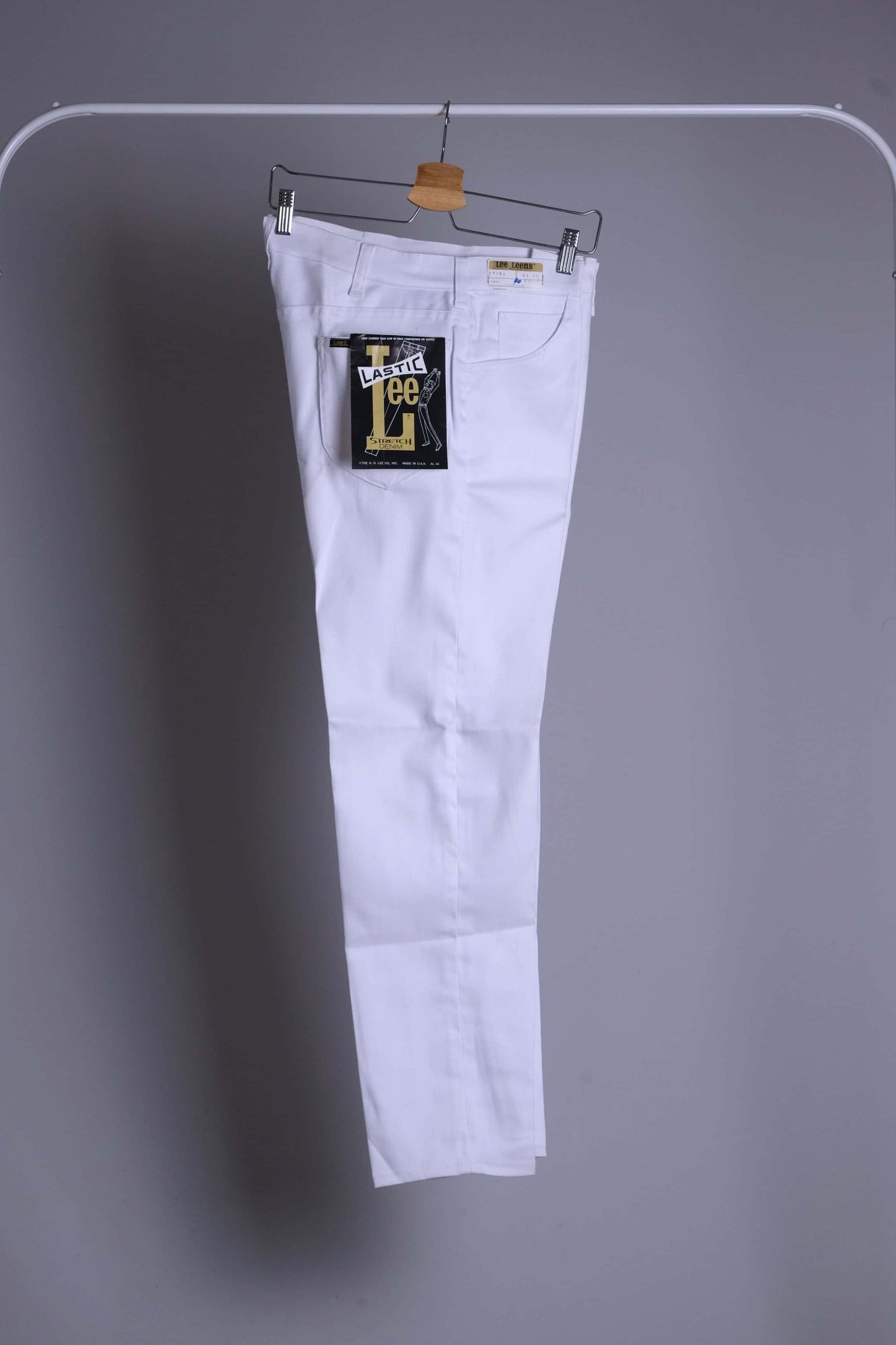 NOS 1970s Lee Leens Lastic Stretch denim trousers in crisp white, front view, hanging with the iconic black and gold 'Lastic Lee' product tag visible on the back pocket.