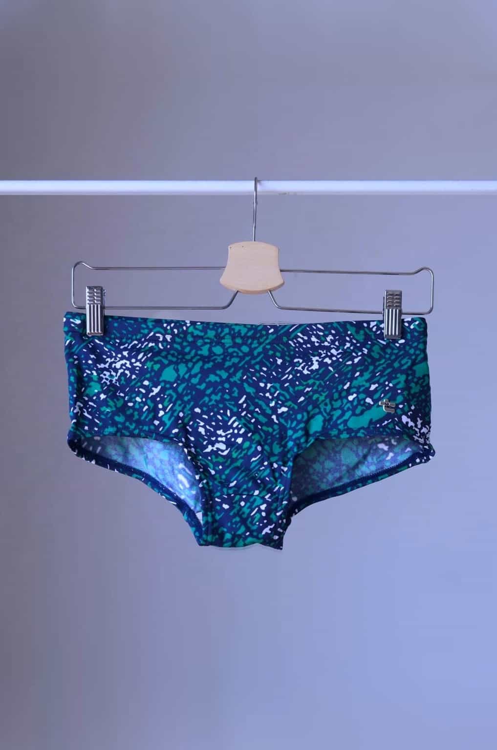 Green swim briefs with an abstract leopard print pattern, hanging on a wooden hanger. The pattern includes shades of blue, green, and white.