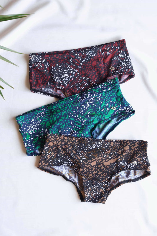 Three pairs of vintage TROPIC Mica swim briefs stacked on top of each other.
 The briefs are in different colors, including maroon, green, and brown, and feature an abstract, leopard-print pattern.