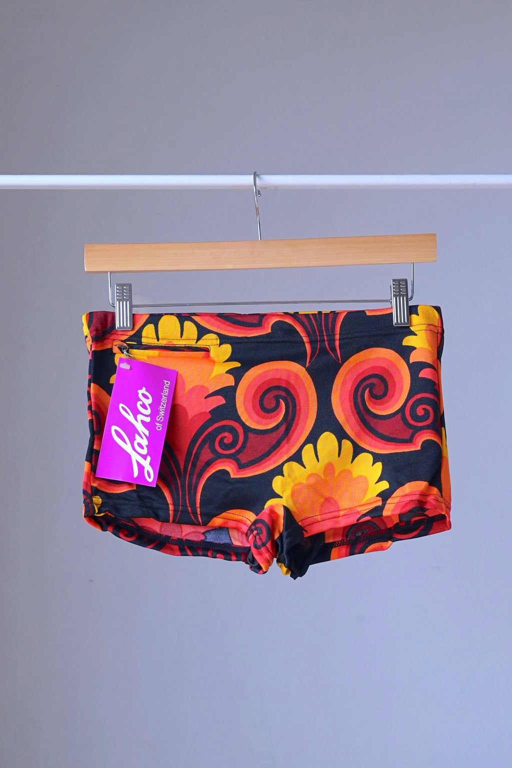 LAHCO vintage 60s square-cut swim trunks in red, yellow, and black psychedelic print with original tag, hanging on a wooden hanger.
