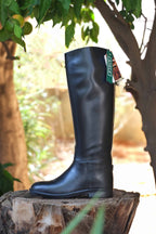 A black rubber horseback riding boot with a leather-look finish, displayed on a wooden stump with a green tag attached to it.